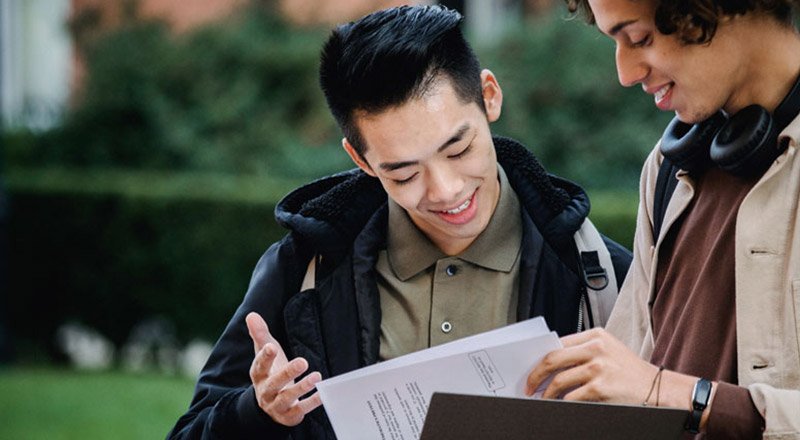Two students outdoors, smiling as they review a document together.