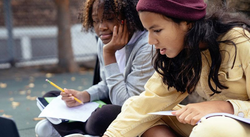 Two young women study outside on a bench, one writing in a notebook while the other rests her head on her hand and watches.