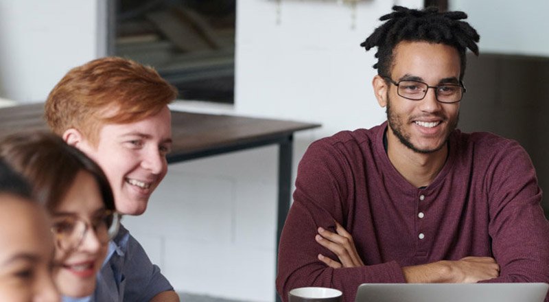 Group of students collaborating at a table, smiling, with a laptop in view in a classroom.