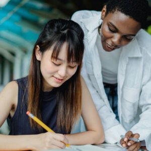 Two students studying outdoors: a woman writes in a notebook while a man leans over to help and smiles.