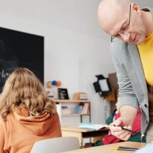 Teacher assists a student at a desk in a bright classroom; chalkboard with drawings in the background.