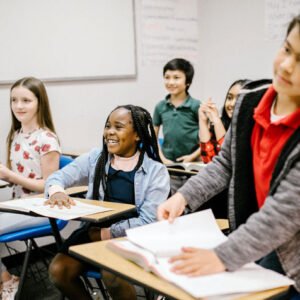 Diverse group of students in a classroom, smiling and engaged at their desks as a student at the front organizes papers.