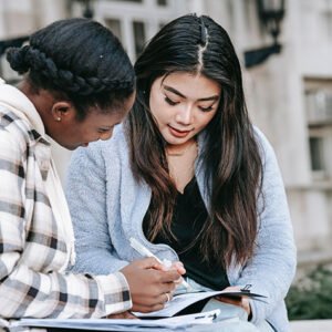 Two women sit on a bench outdoors, looking over papers and studying together.