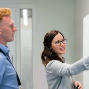 Woman in glasses writes on a whiteboard as a male colleague in a blue shirt watches during a meeting in a modern office.