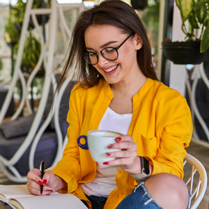 Woman with glasses in a yellow jacket writing in a notebook while smiling and holding a cup in a cozy cafe garden setting.