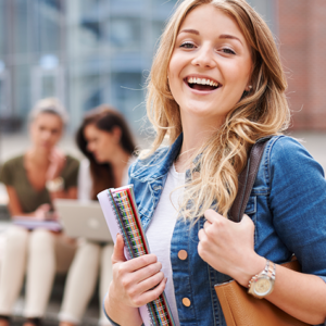 Young woman smiling outdoors on a campus, holding notebooks and a bag, with two classmates on steps in the background.