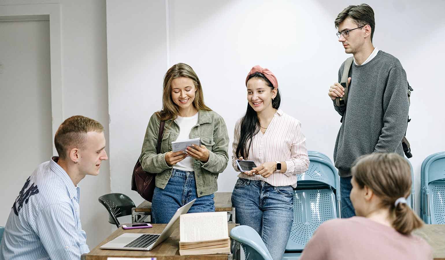 Group of students gathered around a table in a classroom, one seated with a laptop and open book, others standing and smiling with phones in hand.