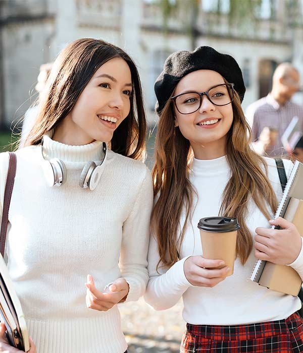 Two smiling young women walking outdoors, one with headphones around her neck, the other holding a coffee cup and notebooks.