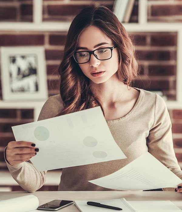 Woman wearing glasses reads printed documents at a desk, concentrating on the papers.