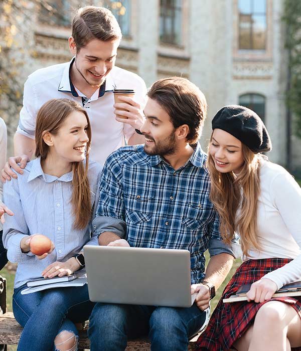 Four friends outdoors, gathered around a laptop, smiling at the screen; one holds a coffee cup.