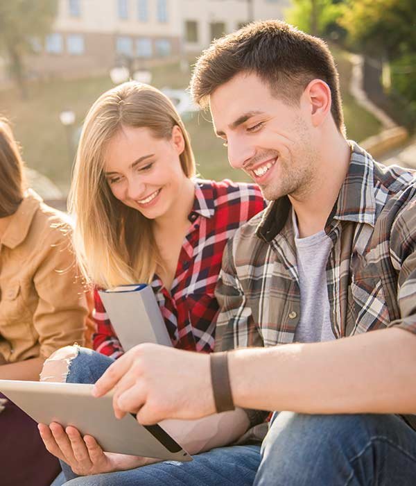 Two friends smile while looking at a tablet outdoors on a sunny day.