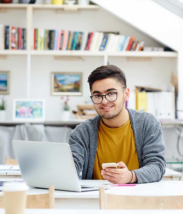 Young man with glasses sits at a bright study desk, using a laptop and holding a smartphone.
